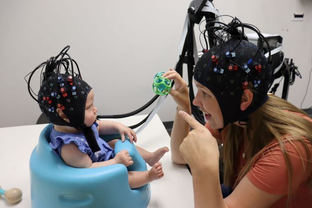Infant and mother in neuroimaging headgear playing at a table top.